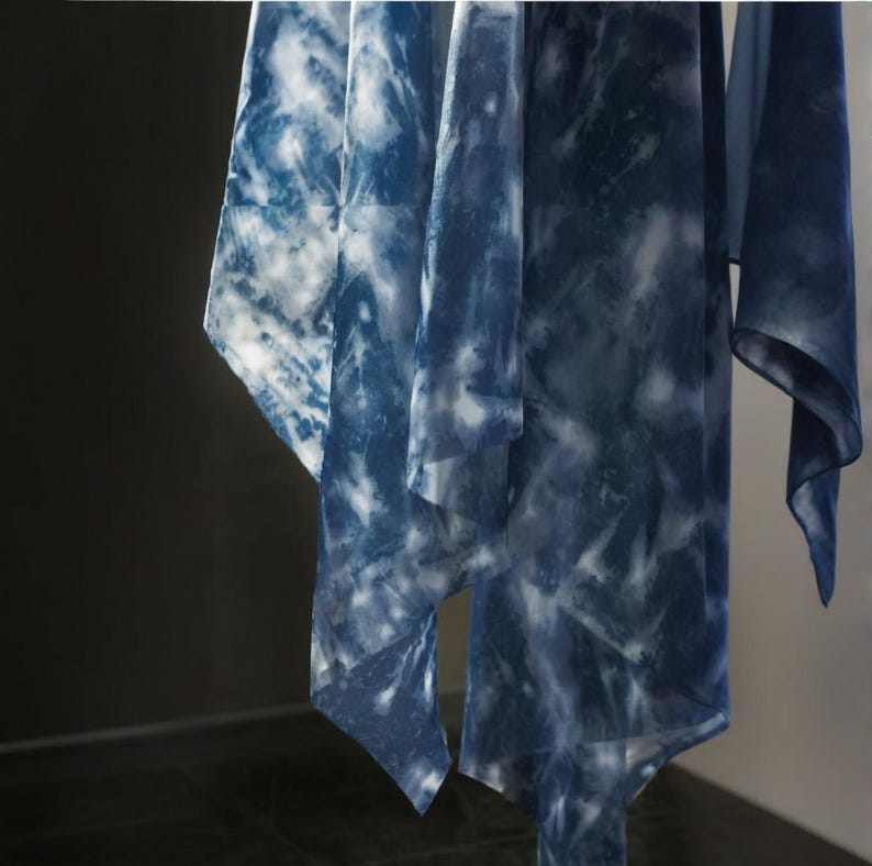May include: Several flowing, sheer fabrics hang from a rod. The fabrics feature a tie-dye pattern in shades of indigo and white, creating a cloudy, marbled effect. The textiles appear lightweight and airy, suggesting they are scarves or shawls.