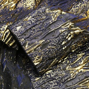 Puede incluir: Una prenda de tela con una base azul marino profundo, una textura arrugada en relieve y un revestimiento de lámina de oro metálico. La superficie muestra un patrón abstracto complejo donde los reflejos dorados contrastan con la tela azul.