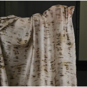 Puede incluir: Bufanda drapeada, blanquecina, con un patr&oacute;n repetido de pinceladas marrones y verde oliva. La tela tiene una textura suave y fluida, y los bordes est&aacute;n ligeramente deshilachados. La bufanda es probablemente de un material ligero.