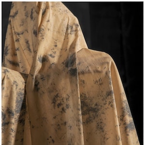 May include: Close-up of tan fabric with dark grey splattered pattern. The fabric is draped over an unseen form, creating folds and shadows. The material appears lightweight and textured, with a natural, organic aesthetic.