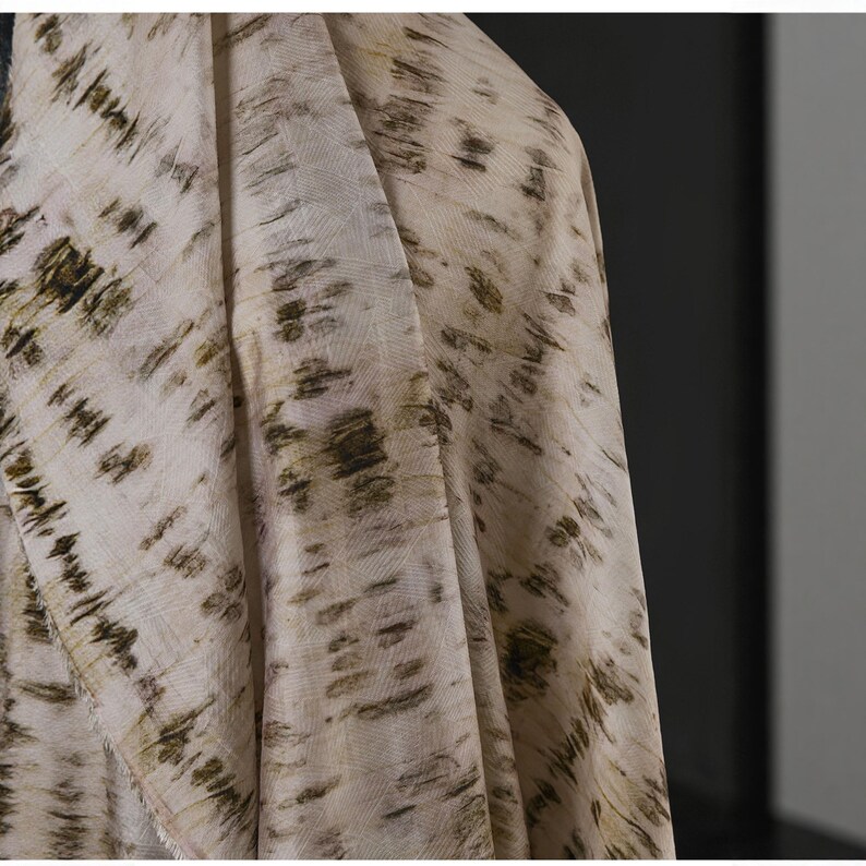 Puede incluir: Primer plano de una tela drapeada con un dise&ntilde;o tie-dye. La tela es principalmente blanquecina con acentos verde oliva oscuro y marr&oacute;n. El patr&oacute;n tiene un aspecto natural y org&aacute;nico, que recuerda a pinceladas o patrones de hojas.