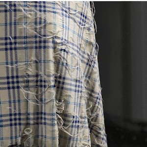 May include: Close-up of a distressed fabric with a blue and white plaid pattern. The fabric has a worn, frayed appearance with intentional rips and tears, creating a textured, vintage aesthetic. The design features a classic grid layout.