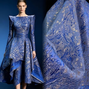 Op de afbeelding: Een jurk met lange mouwen in een diepblauwe stof met een gestructureerd goudkleurig metallic patroon. De jurk heeft gestructureerde schouders, een getailleerde taille en een asymmetrische rok met meerdere overlappende panelen voor een volumineus silhouet.