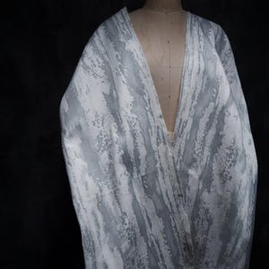 May include: A draped fabric featuring a white and grey abstract pattern with vertical, brushstroke-like textures. The material is displayed on a mannequin, showing a deep V-neckline drape that highlights the layered, textured surface of the textile.