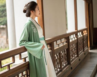 May include: A traditional Chinese Hanfu dress. The dress has a long, flowing white skirt, a white top with floral embroidery, and a light green overcoat. The sleeves are long and flowing. The dress is worn with a matching green belt.