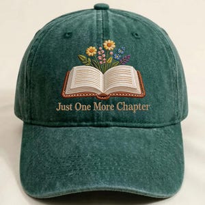 May include: A green baseball cap with an embroidered design featuring an open book with flowers and the text "Just One More Chapter". The cap has a curved brim and a fabric strap closure.