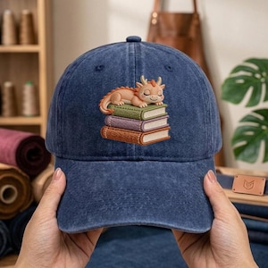 May include: A navy blue baseball cap with an embroidered design of a sleeping dragon on a stack of books. The dragon is light brown with orange accents. The books are green, purple, and brown.