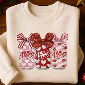 May include: Cream-colored sweatshirt featuring three Dr. Pepper cans with Valentine's Day designs. Each can has a bow and is decorated with hearts, XOXO, or lips. The Dr. Pepper logo is visible on each can.