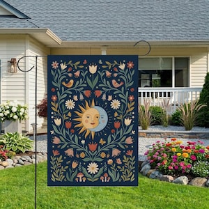 May include: A navy blue garden flag featuring a sun and moon design with floral and bird accents. The sun and moon have smiling faces. The flag is decorated with flowers, butterflies, and mushrooms. The flag is displayed on a black metal stand in a front yard.