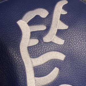 May include: Close-up of a navy blue leather item with a textured surface. The item features a large, stylized, embroidered design in a light gray color. The design appears to be a monogram or a logo.