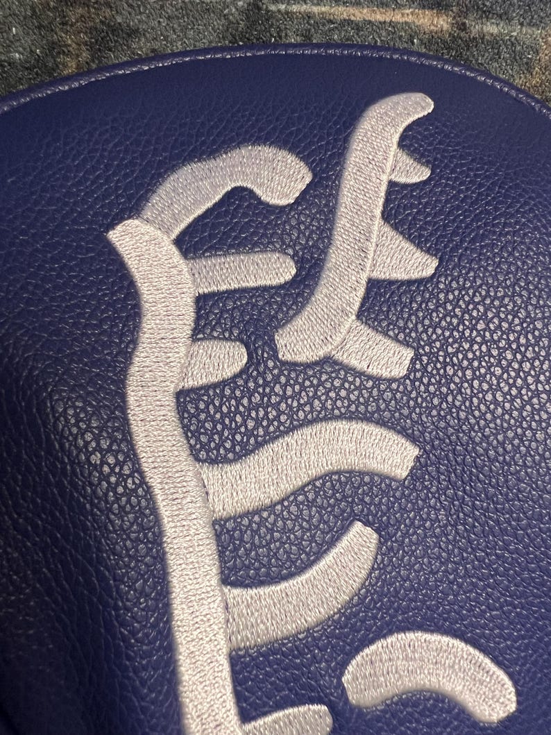 May include: Close-up of a navy blue leather item with a textured surface. The item features a large, stylized, embroidered design in a light gray color. The design appears to be a monogram or a logo.