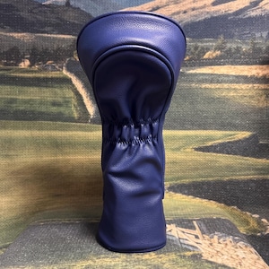 May include: A navy blue golf club head cover. The cover is made of a smooth, leather-like material. The top portion is rounded, and the bottom is gathered with elastic. The background is a landscape scene.