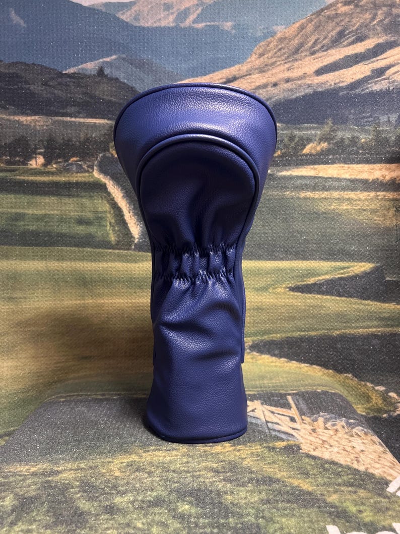 May include: A navy blue golf club head cover. The cover is made of a smooth, leather-like material. The top portion is rounded, and the bottom is gathered with elastic. The background is a landscape scene.