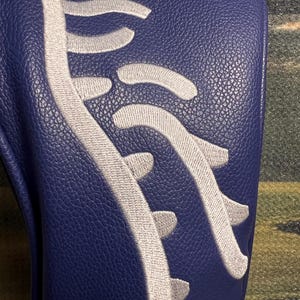 May include: A navy blue golf club head cover with a white embroidered design resembling a spine. The cover has a textured surface and is likely made of leather or a similar material. The design is centered on the cover.