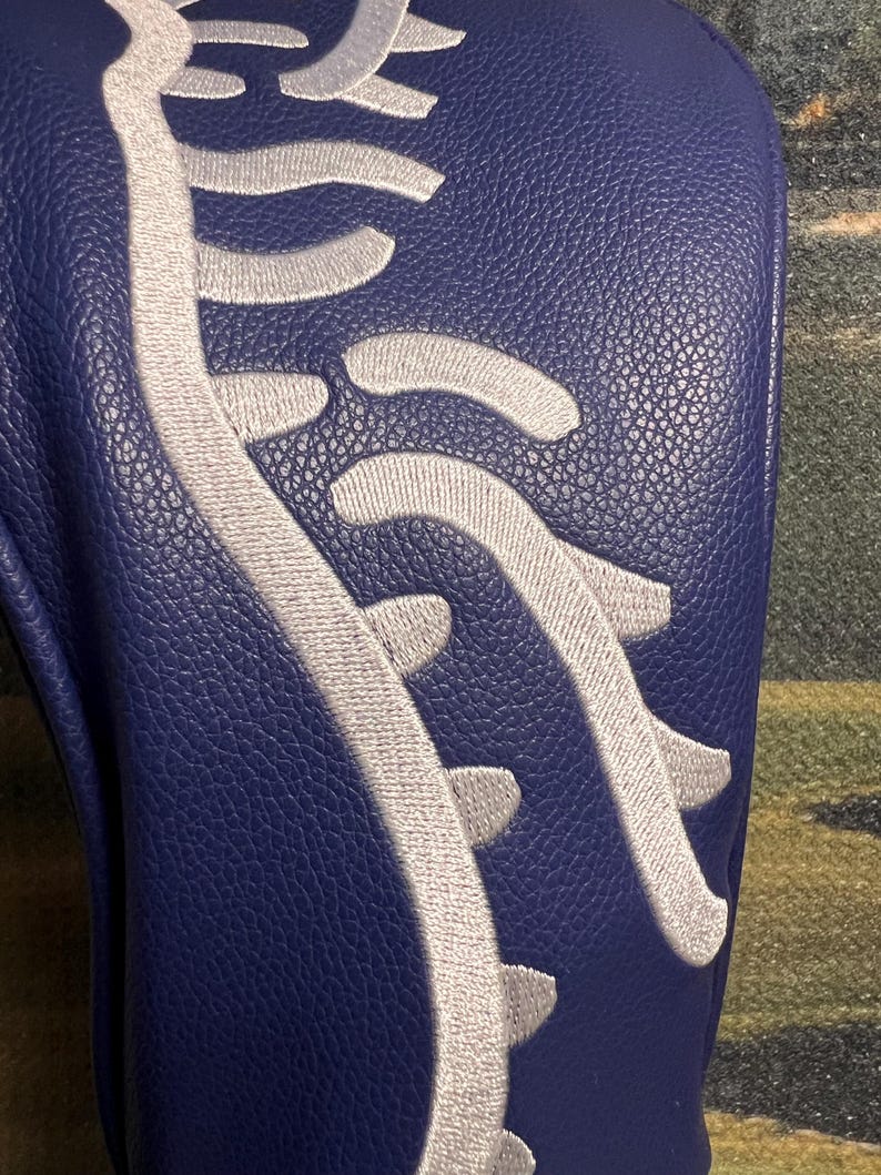 May include: A navy blue golf club head cover with a white embroidered design resembling a spine. The cover has a textured surface and is likely made of leather or a similar material. The design is centered on the cover.