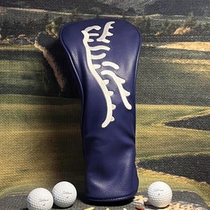 May include: A navy blue golf club head cover with a white abstract design. Three Titleist golf balls and several golf tees are arranged around the cover. The golf balls are white with black text and the tees are various colors.