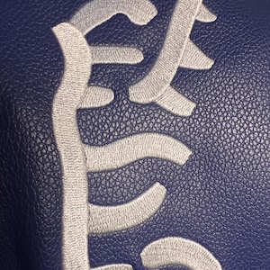 May include: A close-up of a navy blue leather item with a white embroidered design. The design is an abstract, stylized script. The leather has a textured, pebbled appearance.