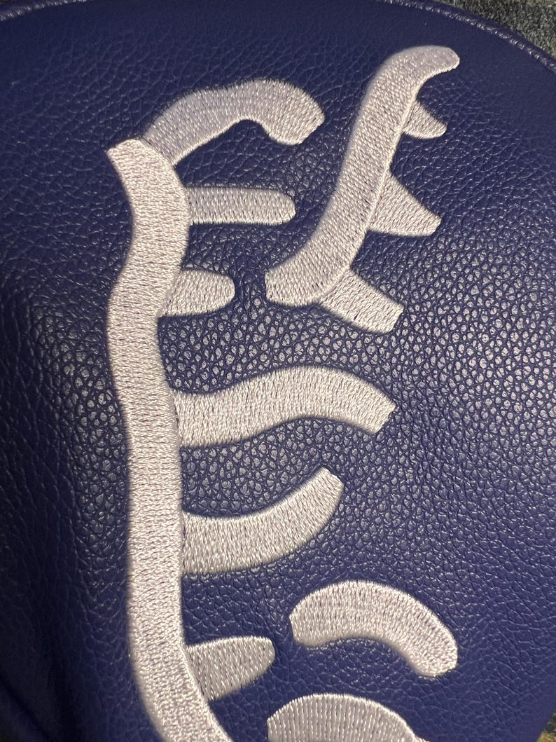 May include: A close-up of a navy blue leather item with a white embroidered design. The design is an abstract, stylized script. The leather has a textured, pebbled appearance.