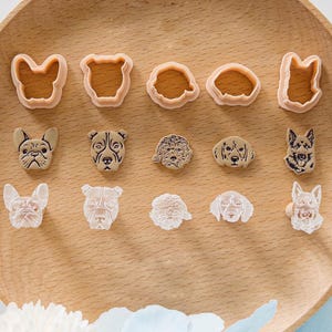 May include: A collection of dog-shaped cookie cutters and decorative dog face embellishments in various breeds. The cutters are light orange, and the dog faces are beige and white, arranged on a wooden surface.