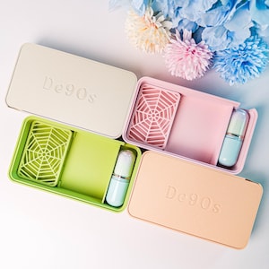 May include: Four rectangular cosmetic cases in pastel colors: beige, pink, green, and peach. Each case contains a small brush and a web-shaped insert. The cases are labeled "De90s" and are arranged with colorful artificial flowers in the background.