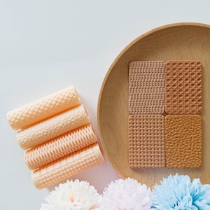 May include: A wooden plate holds four textured, rectangular coasters in shades of tan and brown.  To the left, several cylindrical, textured objects in a light peach color are stacked.  Floral accents in white, pink, and blue are at the bottom.