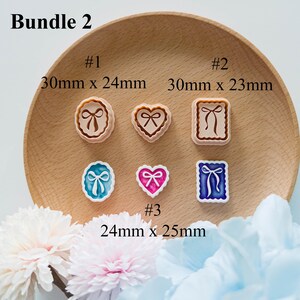 May include: A collection of six decorative stamps in various shapes, including round, heart, and square, each featuring a bow design. The stamps are in shades of brown, pink, blue, and white. Measurements are provided in millimeters.
