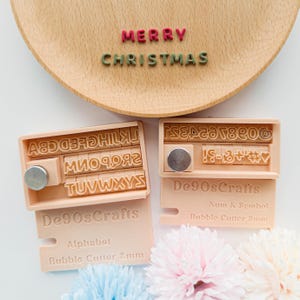 May include: A wooden tray with the words "MERRY CHRISTMAS" in red and green letters. Two sets of alphabet and number stamps are displayed. The stamps are labeled "De90sCrafts" and "Alphabet Bubble Cutter 8mm".