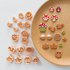 May include: A collection of light peach plastic cookie cutters and matching finished clay charms arranged on a wooden plate. The shapes include mushrooms, squirrels, owls, leaves, mugs, a round door, and a barn, featuring detailed embossed designs.
