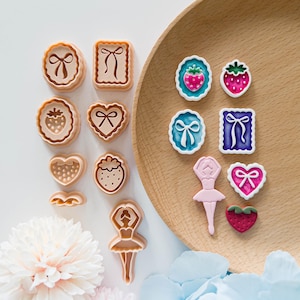 May include: A collection of cookie cutters and decorative embellishments in various shapes, including bows, strawberries, hearts, and a ballerina. The cutters are beige, while the embellishments feature pink, blue, and green accents. The items are arranged on a wooden plate and white surface.