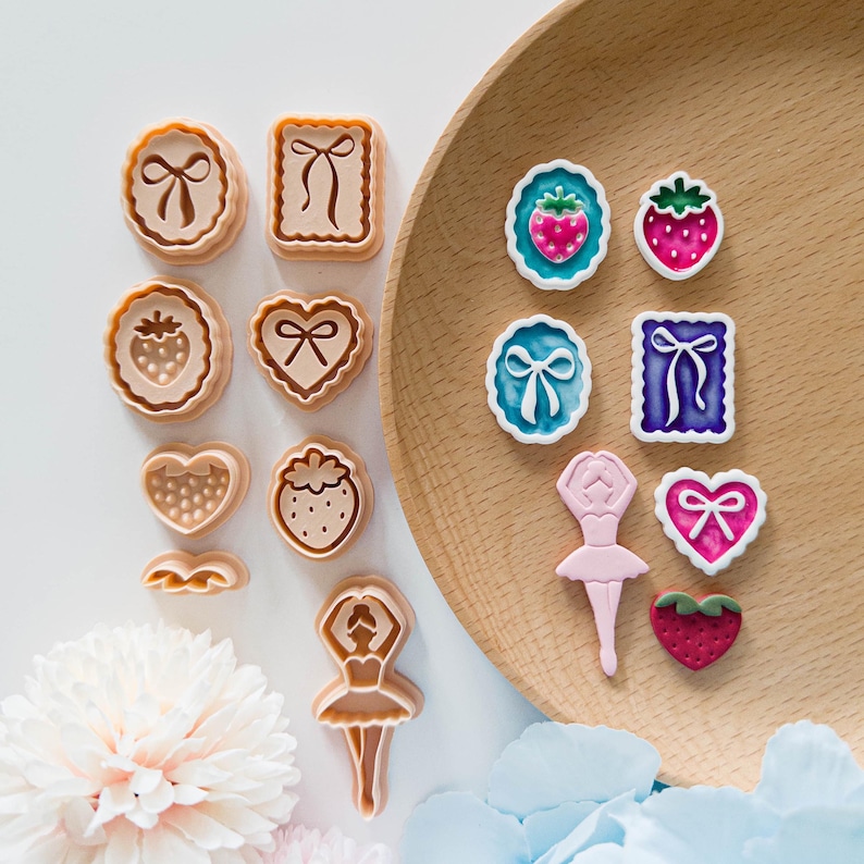 May include: A collection of cookie cutters and decorative embellishments in various shapes, including bows, strawberries, hearts, and a ballerina. The cutters are beige, while the embellishments feature pink, blue, and green accents. The items are arranged on a wooden plate and white surface.