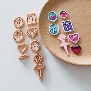 May include: A collection of cookie cutters and decorative embellishments. The cutters are in various shapes, including hearts, circles, and squares, with bow and strawberry designs. Embellishments include strawberries, bows, and a ballerina. The items are in shades of pink, white, and blue.