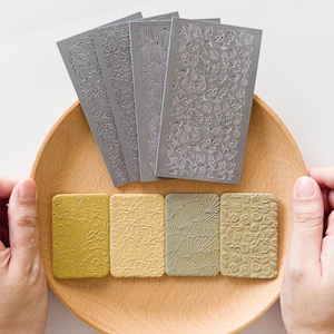 May include: A wooden tray holds several decorative items. There are four rectangular, textured blocks in shades of yellow and green, and five gray, embossed metal sheets with floral and botanical designs. The items are arranged on a light wooden tray.