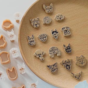 May include: A collection of dog-shaped cookie cutters and decorative dog-face embellishments. The cookie cutters are light brown, and the dog faces are beige with black outlines. The items are arranged on a wooden tray and a white surface.