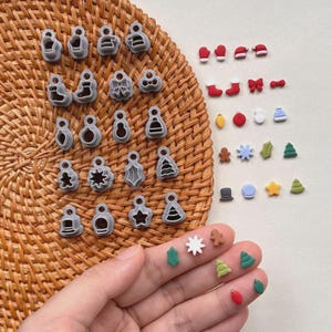 May include: A collection of Christmas-themed cookie cutters and decorative embellishments. The cutters are gray plastic in various shapes, including ornaments, bells, and stars. The embellishments are small, colorful, and include mittens, hats, and Christmas trees.