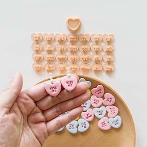 May include: A pair of pink heart-shaped earrings with the text "KISS ME" and pearl accents are held in a hand. Scattered around are various pastel-colored heart-shaped candies with phrases like "CUTIE PIE" and "LOVE ME".