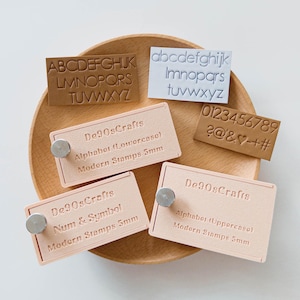 May include: A collection of modern stamps, including uppercase and lowercase alphabets, numbers, and symbols. The stamps are displayed on a wooden tray. The stamps are in various colors, including tan and white. The text "De90sCrafts" is visible on the stamp holders.