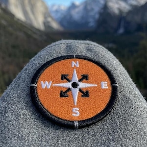 May include: A gray fabric item with an embroidered compass patch. The compass is orange with black and white details, featuring directional indicators. The background shows a scenic mountain landscape under a clear blue sky.