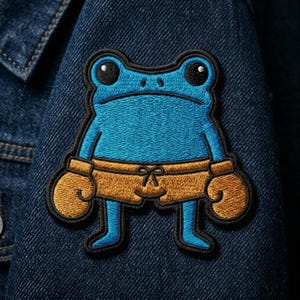 May include: Embroidered patch featuring a blue frog wearing boxing gloves and shorts. The frog has a sad expression and is outlined in black. The patch is attached to a denim jacket.