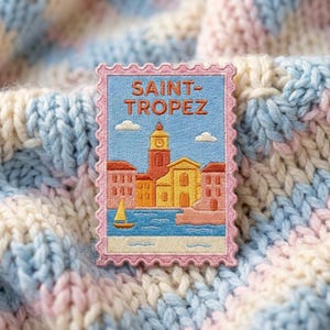 May include: A rectangular embroidered patch shaped like a postage stamp with a scalloped pink border. The design features a light blue sky with white clouds, a yellow sailboat on blue water, and a coastal town with orange and yellow buildings.