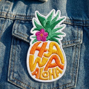 May include: An embroidered patch shaped like a pineapple with green leaves and a pink flower at the top. The body of the pineapple features the orange and yellow text HAWAII ALOHA integrated into the design, all outlined in white thread.