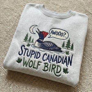 May include: A light gray sweatshirt with a crew neck featuring an embroidered design. The design includes a loon wearing a red hat, a crescent moon, and the text "Awooo?" in a speech bubble. Below, the text reads "Stupid Canadian Wolf Bird."