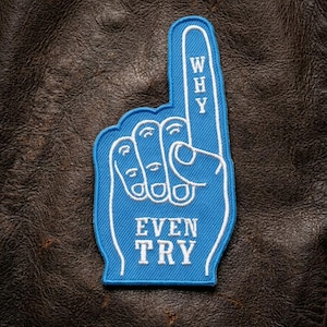 May include: A blue foam finger patch with white trim and the words "WHY" and "EVEN TRY" in white lettering. The patch is on a dark brown leather-like surface.