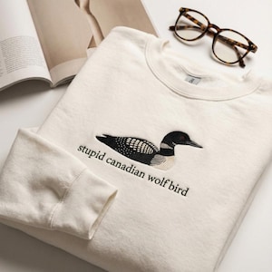 May include: Cream-colored sweatshirt with an embroidered loon design in black and white, and the text "stupid canadian wolf bird". Tortoiseshell glasses rest on the surface.