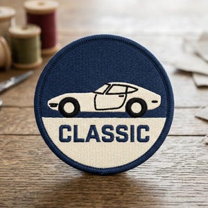 May include: A navy blue and cream-colored embroidered patch featuring a classic car design. The car is white, with black wheels and trim. The word "CLASSIC" is embroidered in white on a cream-colored background.