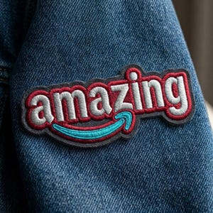 May include: Embroidered patch with the word "amazing" in white, outlined in red, and a gray border. A blue curved arrow is below the word. The patch is sewn onto a blue denim jacket.