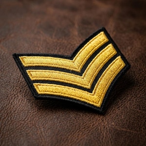 May include: Embroidered patch featuring a gold chevron design with three stripes on a black background. The patch is set against a brown leather-like surface. This patch is likely used for military or uniform insignia.