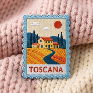 May include: A rectangular embroidered patch shaped like a postage stamp with a light blue scalloped border. The design depicts a Tuscan landscape with a yellow house, rolling orange fields, dark blue cypress trees, and a red sun. The word TOSCANA is stitched in red at the bottom.