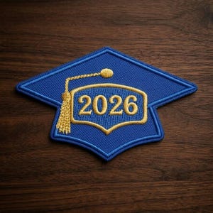May include: A blue graduation cap patch with gold embroidery. The patch features the year "2026" in a gold frame, a gold tassel, and a gold button. The patch is on a dark wood surface.