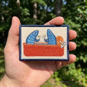 May include: A rectangular embroidered patch featuring two blue fish inside an orange sardine tin. The fish are holding small martini glasses. The patch has a cream-colored background and a dark blue border, with a silver-colored pull tab visible.