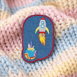 May include: An oval-shaped embroidered patch with a blue denim background and a red border. The design features a silver rocket ship with yellow fins and a bright orange flame, positioned above a colorful planet with blue, yellow, and red swirls.